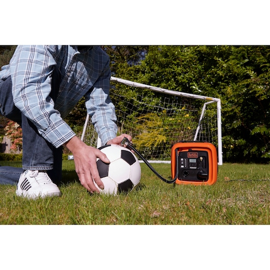 BLACK+DECKER ASI400 portable air compressor inflating a soccer ball on grass near a soccer goal.