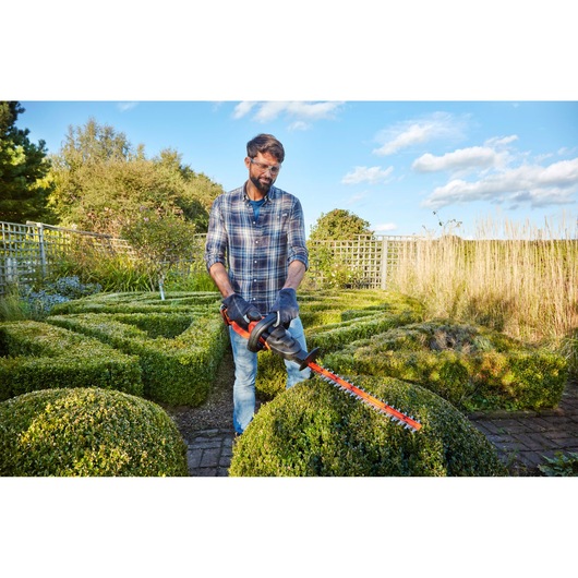 Man using BLACKDECKER hedge trimmer BCASK861D in a garden, trimming green hedges on a sunny day.