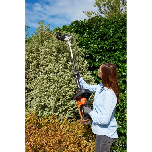 BLACK+DECKER BCASK861D pole hedge trimmer in use trimming tall garden hedge, side view outdoors.