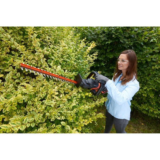 Person using BLACK+DECKER BCASK861D hedge trimmer in garden trimming green bushes.