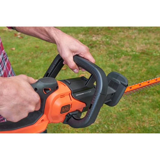 Close-up of BLACK+DECKER BCASK861D hedge trimmer in use, orange and black with visible dual handle grip.