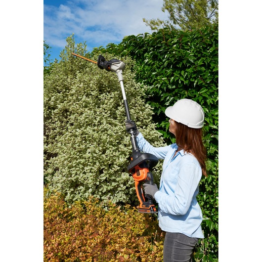 BLACKDECKER BCASK861D pole hedge trimmer in use, woman wearing safety gear trims tall garden hedges.