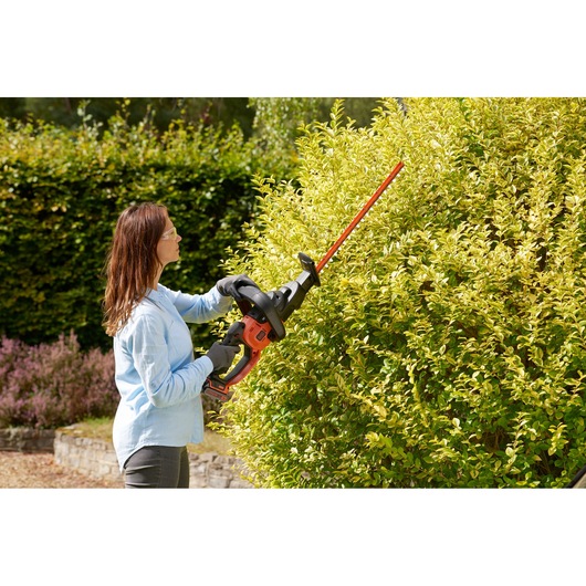 Person using BLACKDECKER BCASK861D hedge trimmer to trim a large bush outdoors.
