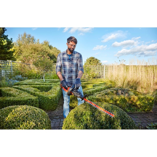 Person trimming bushes in a garden with BLACKDECKER BCASK891D orange hedge trimmer, wearing gloves.