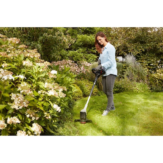 BLACKDECKER BCASK891D cordless grass trimmer used in garden, flower beds visible, outdoor environment.