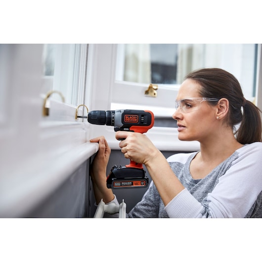 A person using a BLACK+DECKER cordless drill on a window frame, performing a home improvement task indoors. The face of the person is blurred for privacy.