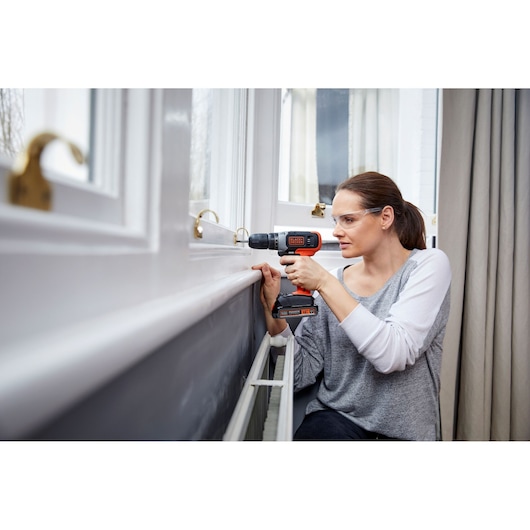 A person using a BLACK+DECKER cordless drill to work on a window frame indoors. The drill is orange and black in color.