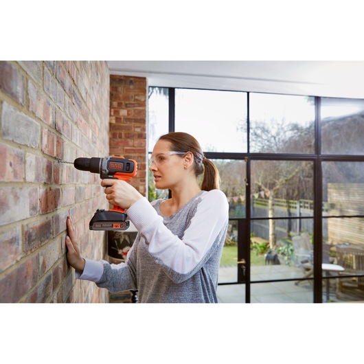 A person using a BLACK+DECKER cordless drill to drill into a brick wall in a bright room with large windows. The individual's face is blurred for privacy.