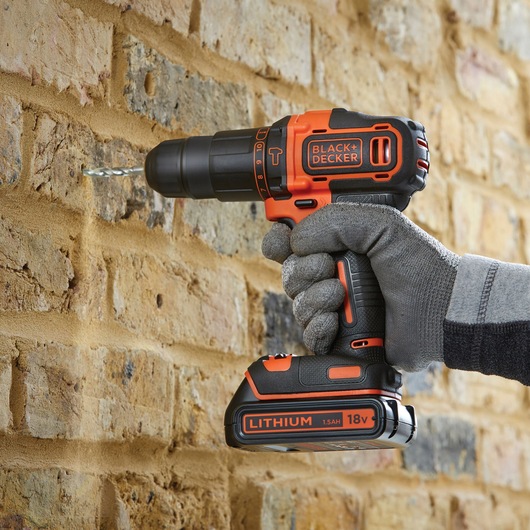 BLACK+DECKER BCD700HTSA cordless drill, black and orange, drilling into brick wall, close-up side view.