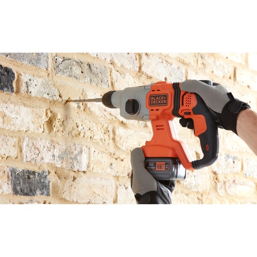 Photo of BLACK+DECKER BCD900M1K cordless hammer drill in use on brick wall, orange and black design, 18V battery visible.