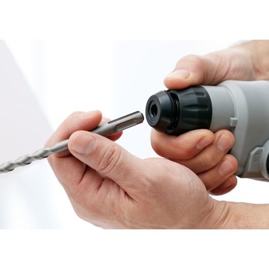 Close-up photo of hands inserting drill bit into BLACKDECKER hammer drill BCD900M1K, white background.