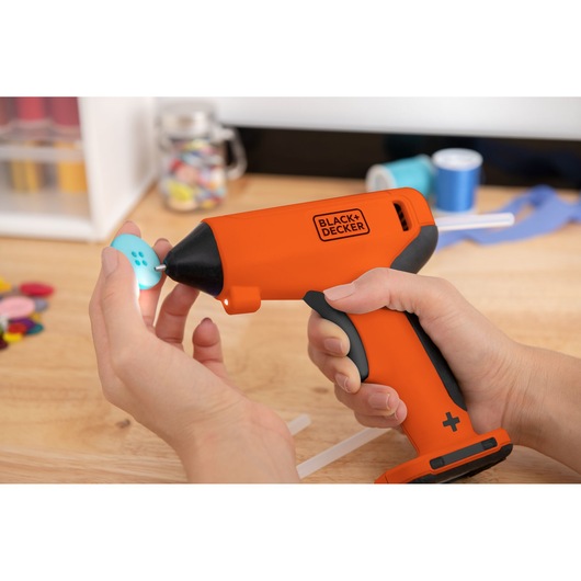 Photo of BLACK+DECKER BCGL115OB cordless glue gun held in hand, applying glue to blue button on wood table.
