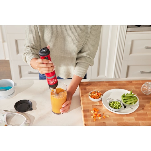 BLACKDECKER BCKM1012KR red hand blender blending orange mixture in tall glass, kitchen counter and vegetables visible.