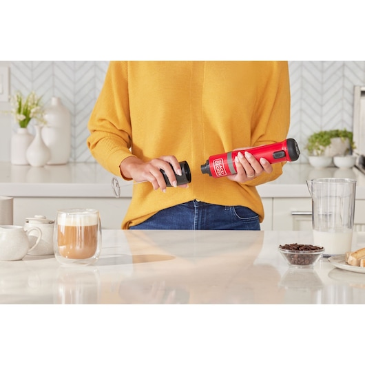 Photo of a person using a BLACKDECKER BCKM1016KSR red handheld milk frother in a kitchen, coffee and milk on counter.