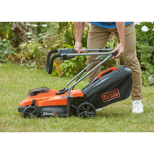 Photo of BLACK+DECKER BCMW3336L2 cordless lawn mower in use on grass, focus on foldable handle and grass collection bag.