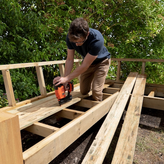 BLACKDECKER BCNG01N cordless nail gun used for outdoor deck construction, worker kneeling on wooden beams.