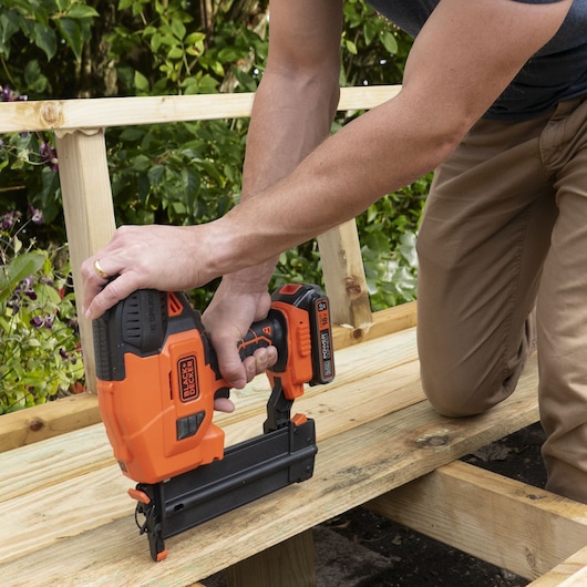 Person using BLACK+DECKER BCNG01N cordless nail gun on wooden outdoor construction.