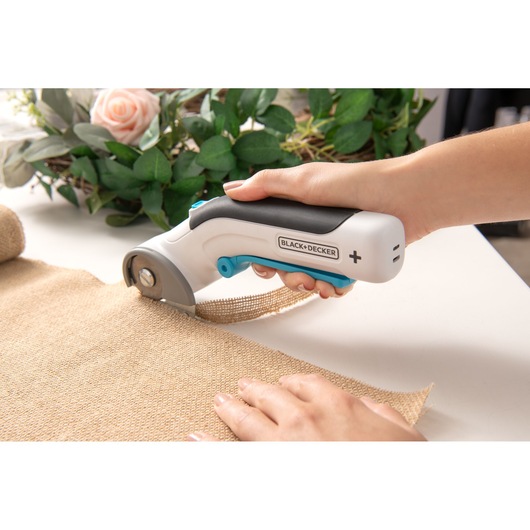 BLACK+DECKER BCRC115 handheld fabric cutter in use, cutting burlap material on a table, close-up view.