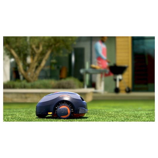 BLACKDECKER BCRMW123 robotic lawn mower on grass, outdoor setting with blurred barbecue in background.