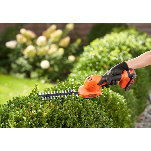 A BLACK+DECKER cordless shrub and grass shear being used to trim a green hedge in a garden. The tool is orange and black, and is held in a gloved hand.