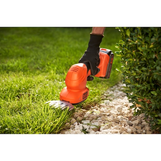 A person uses a BLACK+DECKER cordless grass and shrub shear (model BCSS18B) with an orange body to trim grass along the edge of a stone pathway next to a bush.