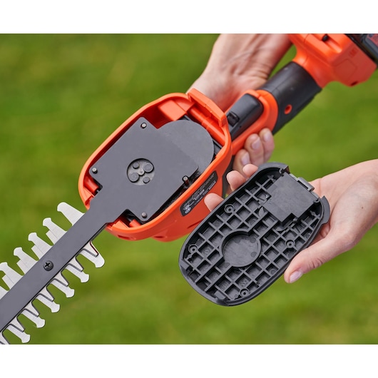 Close-up view of a person holding an orange and black electric garden tool, with the casing opened to show internal parts and blades. The person is holding the removed black plastic cover in their hand.