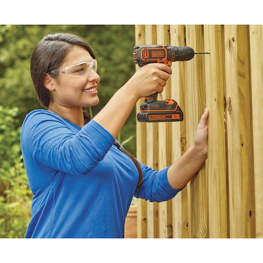 Person using a BLACK+DECKER 18V cordless drill to drill into a wooden fence outdoors. The person's face is obscured.