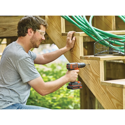 Photo of a person using a BLACK+DECKER BDCDC18BTA cordless drill on a wooden staircase outdoors.