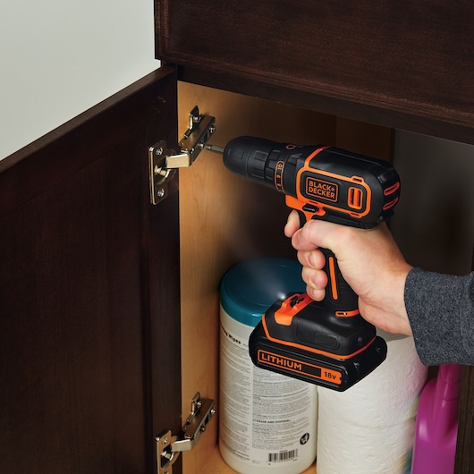 A hand is using a BLACK+DECKER 18V cordless drill to install or adjust a hinge inside a wooden cabinet. Cleaning supplies are visible inside the cabinet.