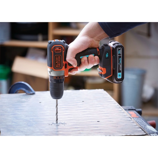 A close-up of a hand holding a BLACK+DECKER cordless drill with an 18V battery, drilling into a metal sheet on a workbench in a workshop.