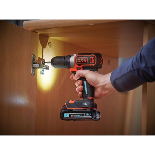 A person using a BLACK+DECKER 18V cordless drill to install a hinge on a wooden cabinet. The drill is equipped with a 'smartech' battery and an LED light illuminating the working area.