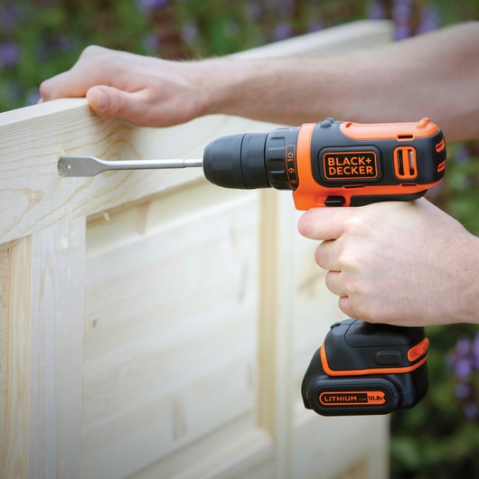 BLACK+DECKER BDCDD12K1B cordless drill in use, orange and black color, drilling wood outdoors.