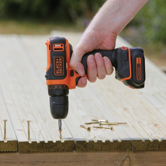 Photo of BLACK+DECKER BDCDD12K1B cordless drill driving a screw into wooden planks, handheld in use outdoors.