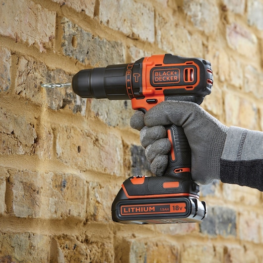 BLACKDECKER BDCHD18BS32 cordless drill in use, drilling into brick wall, lateral view, orange and black design.