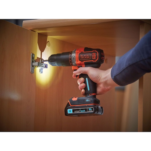 A person using a BLACK+DECKER 18V cordless drill with smart tech, drilling a screw into the hinge of a wooden cabinet door. The drill has an LED light illuminating the work area.