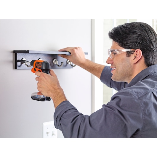Person using a BLACK+DECKER cordless screwdriver to install a coat rack with hooks on a wall.