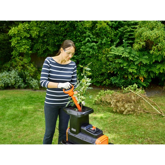 Person using BLACK+DECKER BEGAS5800 garden shredder for branches outdoors, with pile of cut branches nearby.