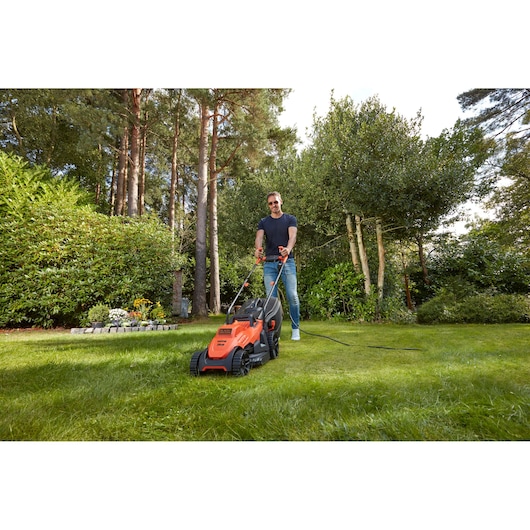 Photo of BLACK+DECKER BEMW461BH orange electric lawn mower in use on a green lawn, surrounded by trees and plants.