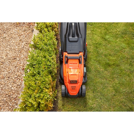 BLACK+DECKER BEMW461BH lawn mower in orange and black trimming grass, top view, close to hedge and gravel path.