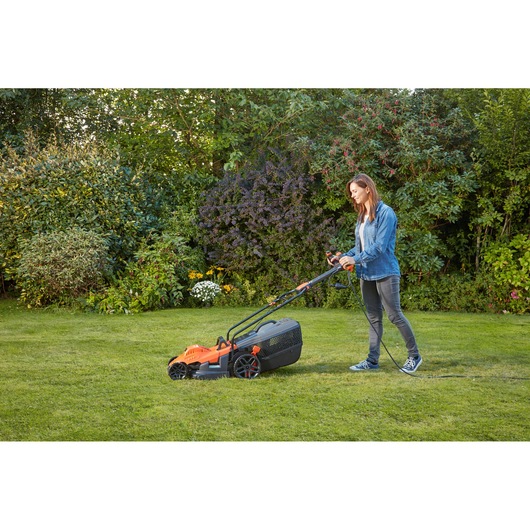 Person using BLACKDECKER BEMW461ES electric lawn mower with orange and black design on grass in garden.
