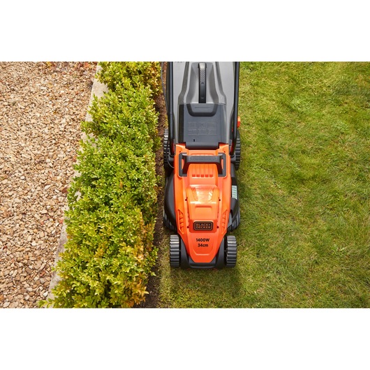 BLACKDECKER BEMW461ES orange lawn mower top view on grass beside hedge and gravel path.