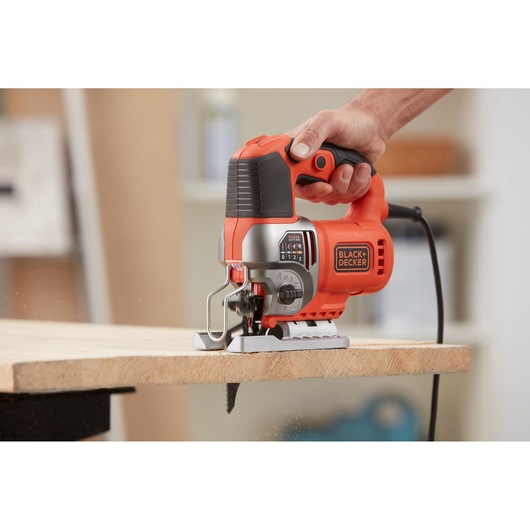 Photo of BLACK+DECKER jigsaw BES610K in use, cutting wood, ergonomic handle, orange and black design.