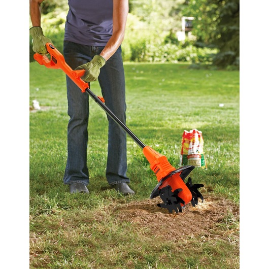 BLACK+DECKER BETL18B cordless garden cultivator in use on a lawn, person wearing gloves operates the orange tool.