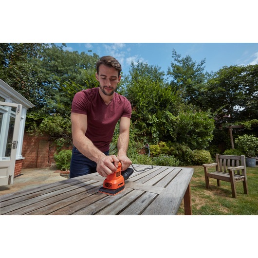 Person using BLACKDECKER BEW200LSA sheet sander on wooden outdoor table in garden setting.