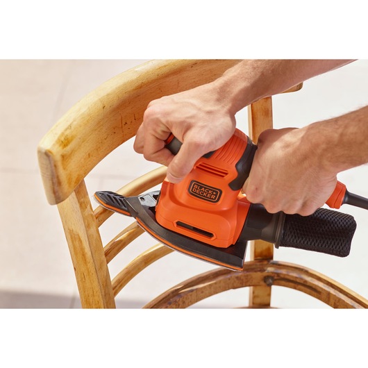 Photo of BLACK+DECKER BEW200LSA sander in use on wooden chair, bright orange and black design.