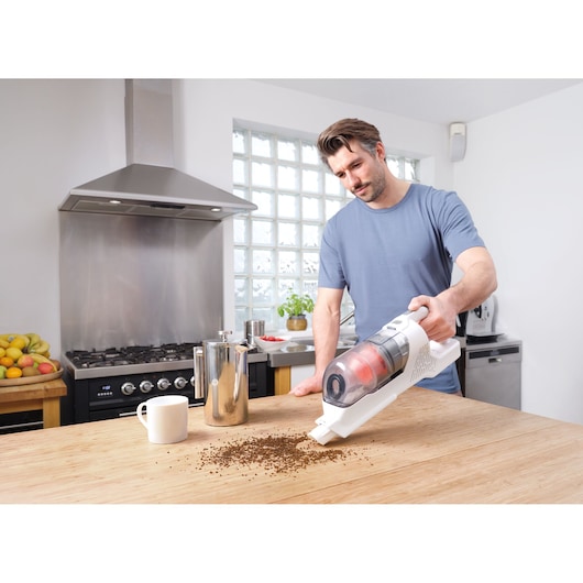 A person using a BLACK+DECKER BHFEA515J handheld vacuum to clean coffee grounds from a wooden kitchen table. The kitchen setting includes a stainless steel stove, a coffee pot, and a mug.