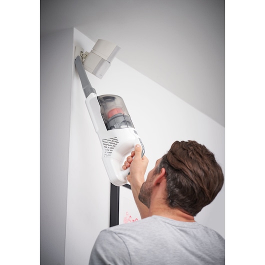 A person uses a BLACK+DECKER cordless handheld vacuum cleaner to clean a high corner of a wall near a ceiling.