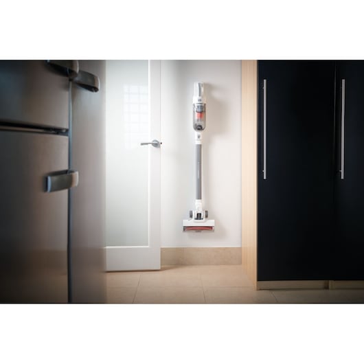 A BLACK+DECKER stick vacuum cleaner is mounted on a white wall in a modern kitchen, positioned between a refrigerator, a cabinet, and a glass door.