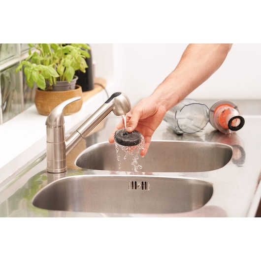 A person rinsing a filter from a vacuum cleaner under a kitchen sink faucet, with vacuum cleaner parts placed on the stainless steel countertop nearby.