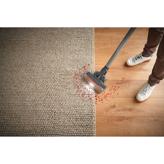 A person uses a BLACK+DECKER cordless vacuum cleaner to clean red crumbs from a light-colored textured carpet and a wooden floor.